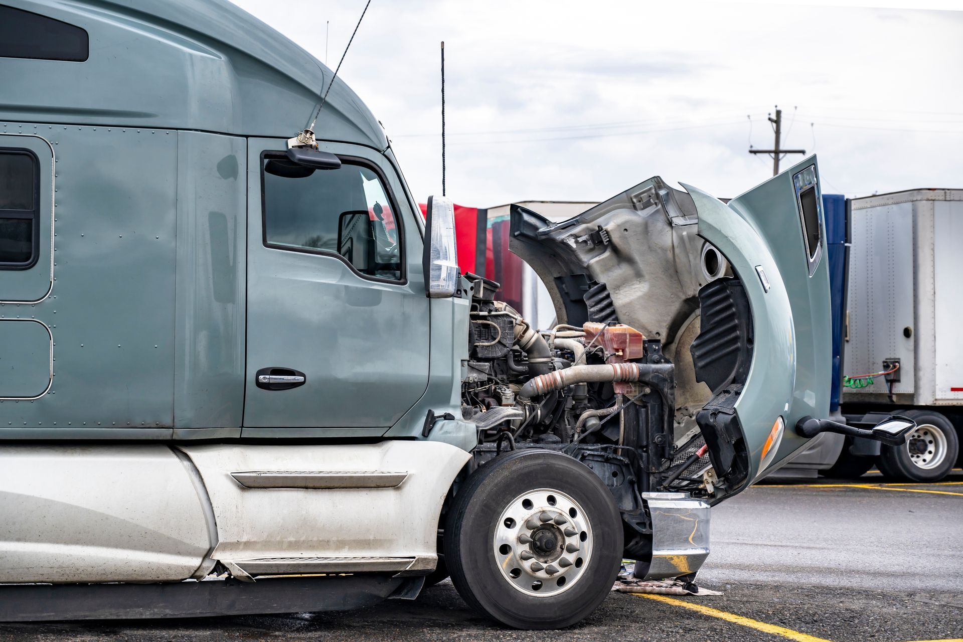 Mechanic in blue uniform inspects truck tires, squatting near wheels; a red forklift is in the background. Semi-truck with open hood; engine visible, parked in lot.