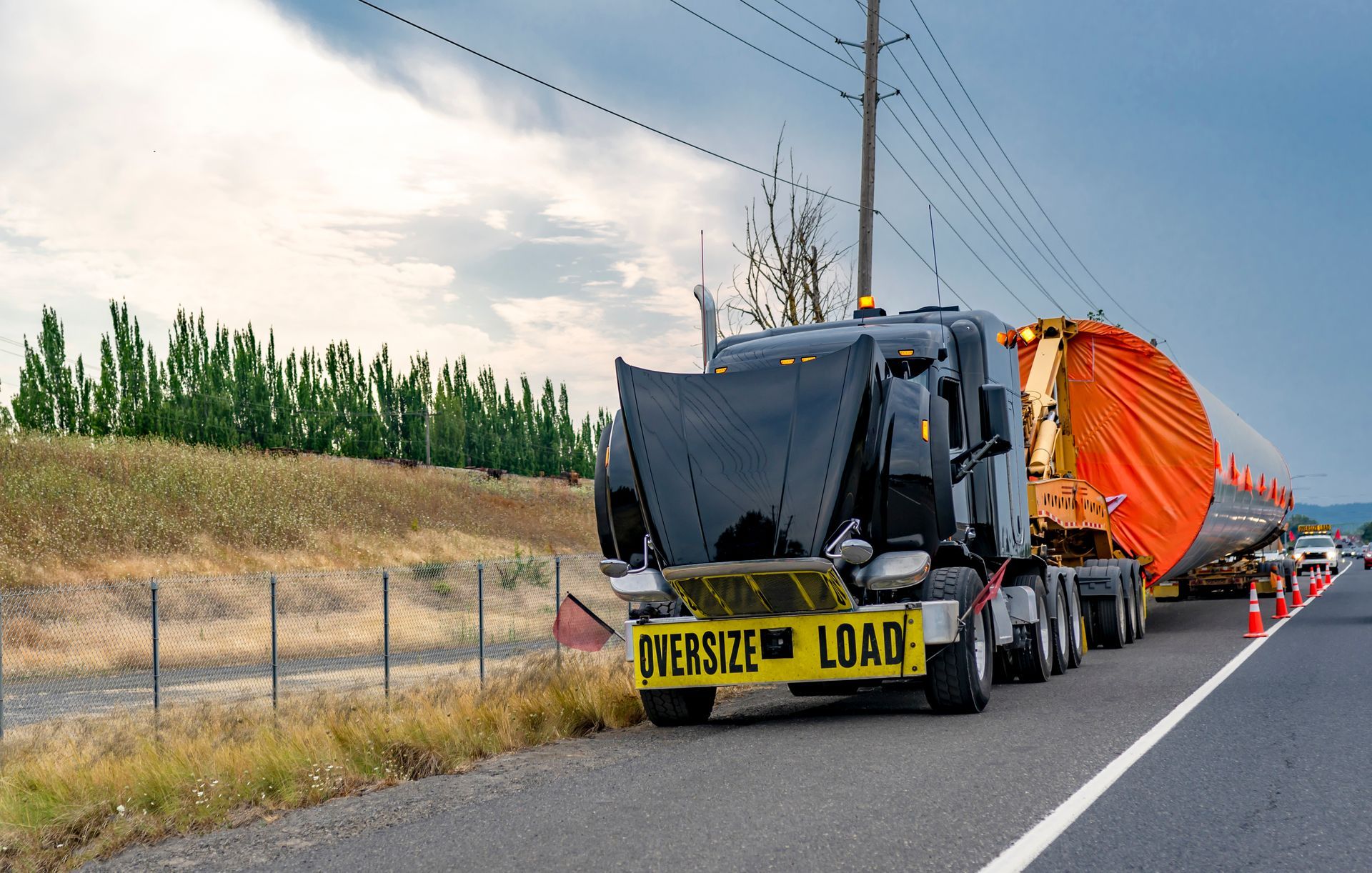 Oversize load truck on a road with a large orange cylindrical object. Black truck, yellow warning sign. Cloudy sky.