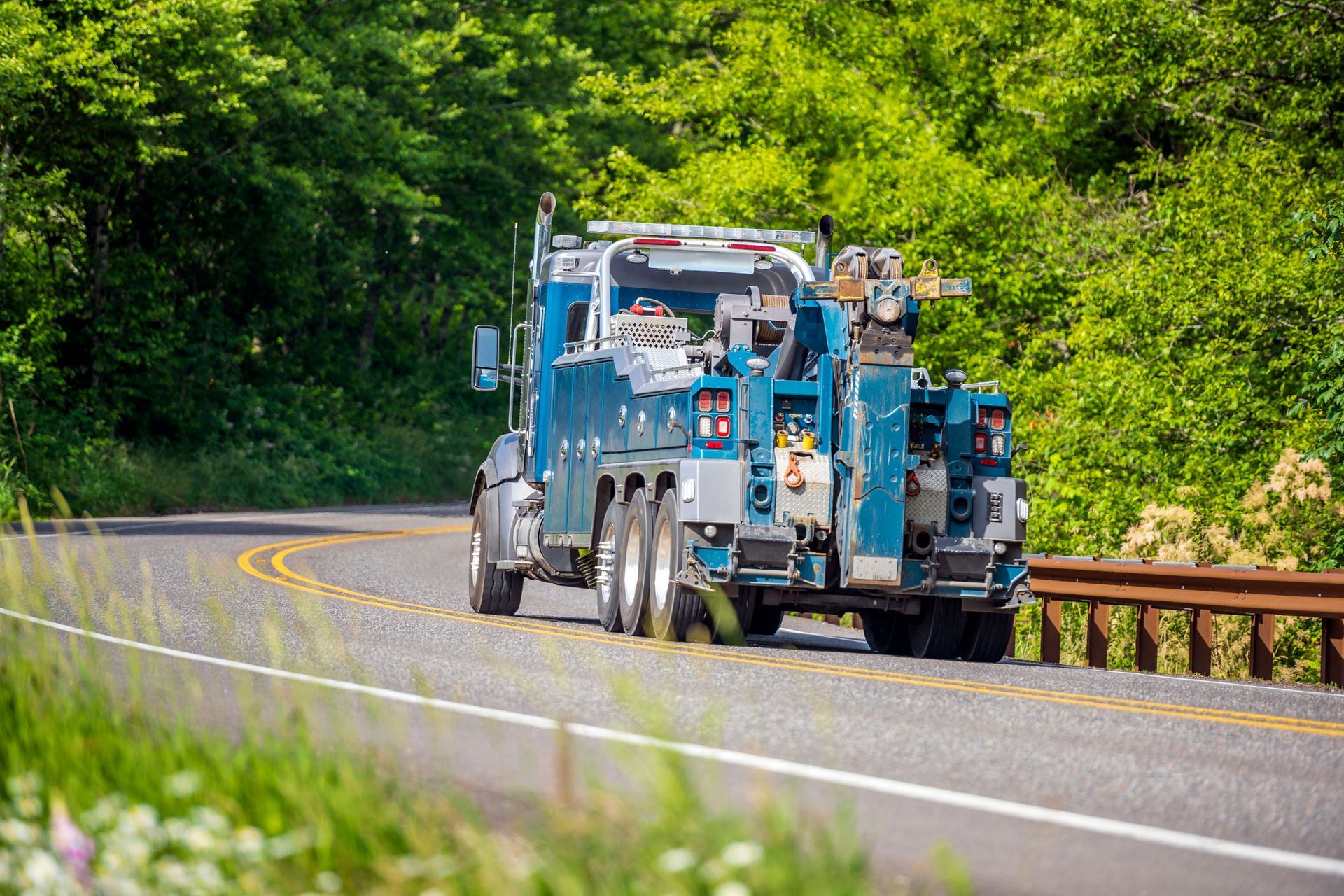 Blue tow truck driving on a curved road, surrounded by green trees and vegetation.