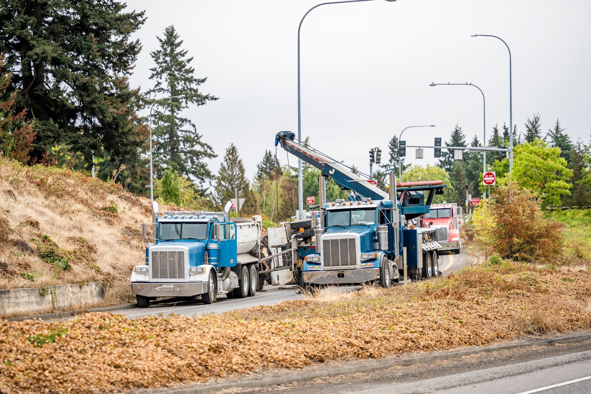 Two blue tow trucks on the side of a road, one lifting a vehicle. Trees and greenery in the background.