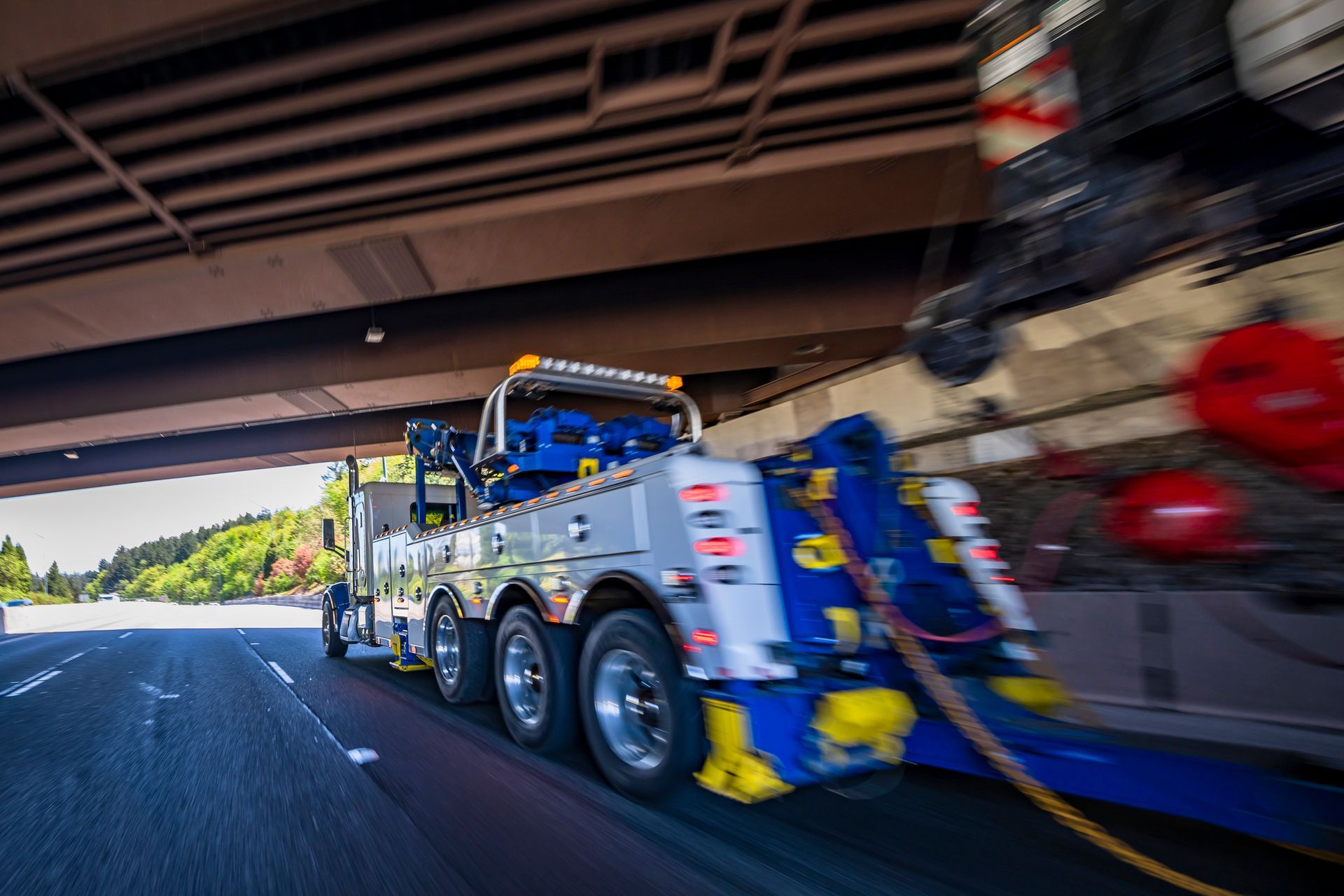 Tow truck towing a car on a highway, seen from a low angle under a bridge.