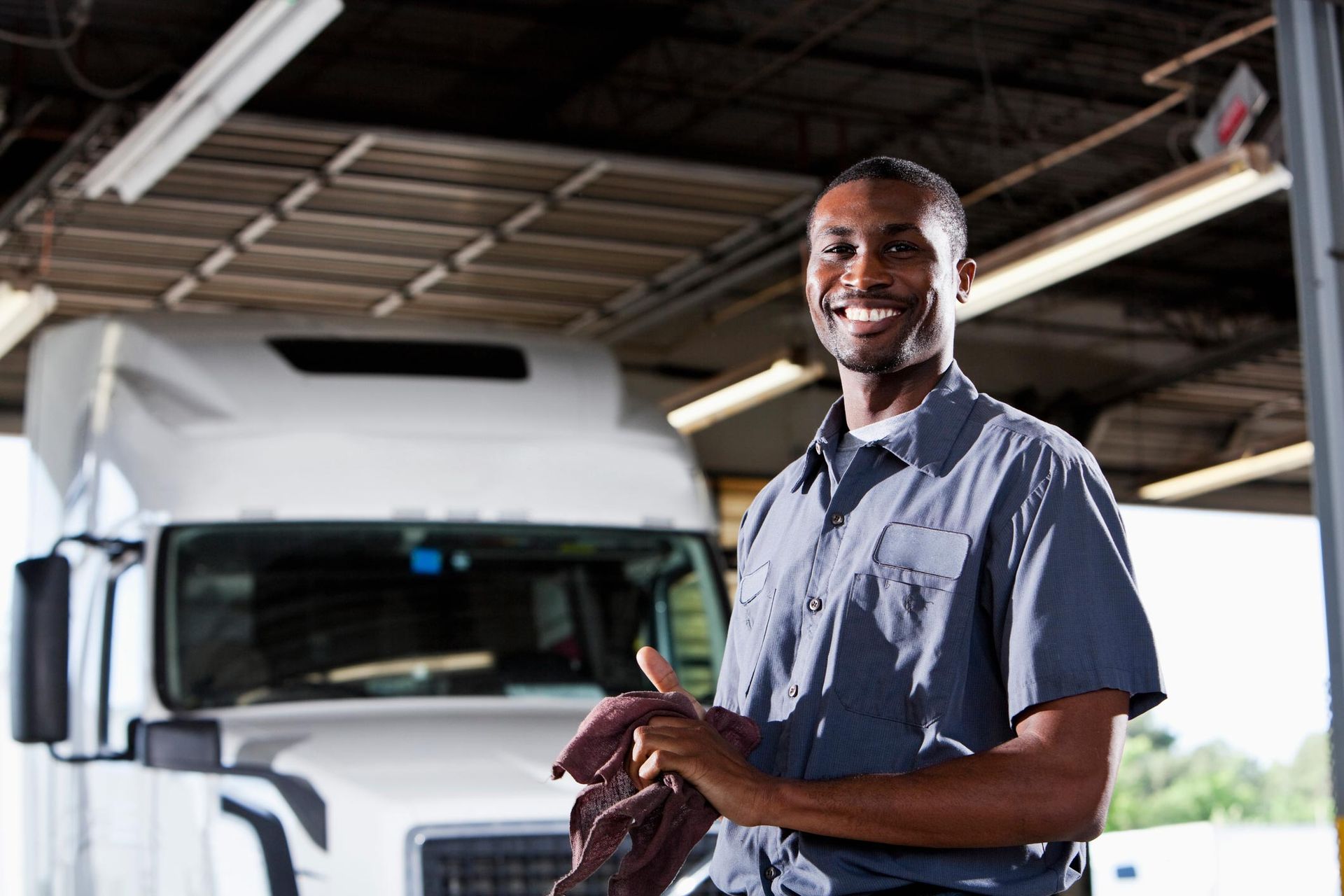 Smiling male truck mechanic in uniform standing in front of semi truck at commercial repair shop.