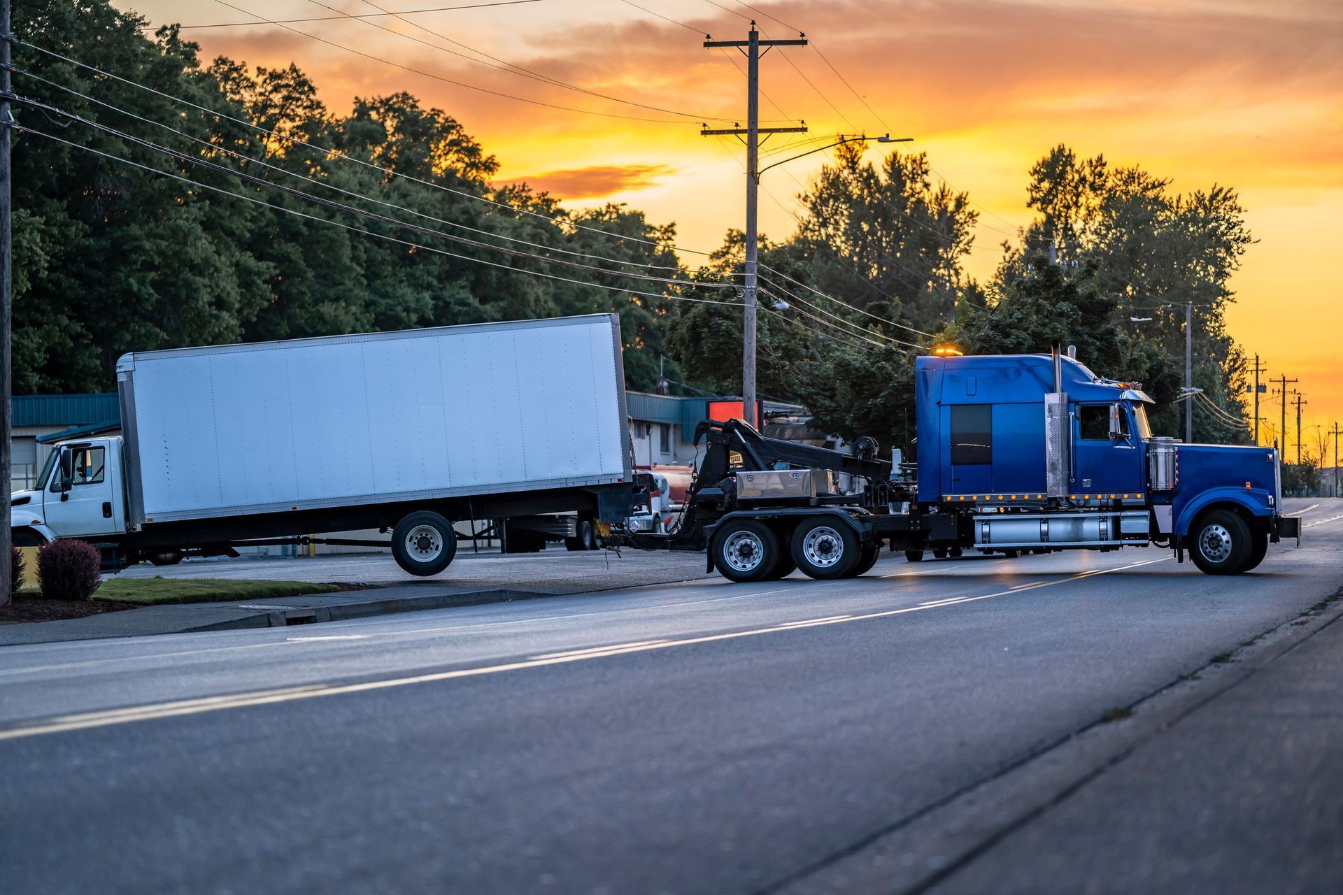 Blue semi-truck towing a white box truck on a road, lit by a sunset.