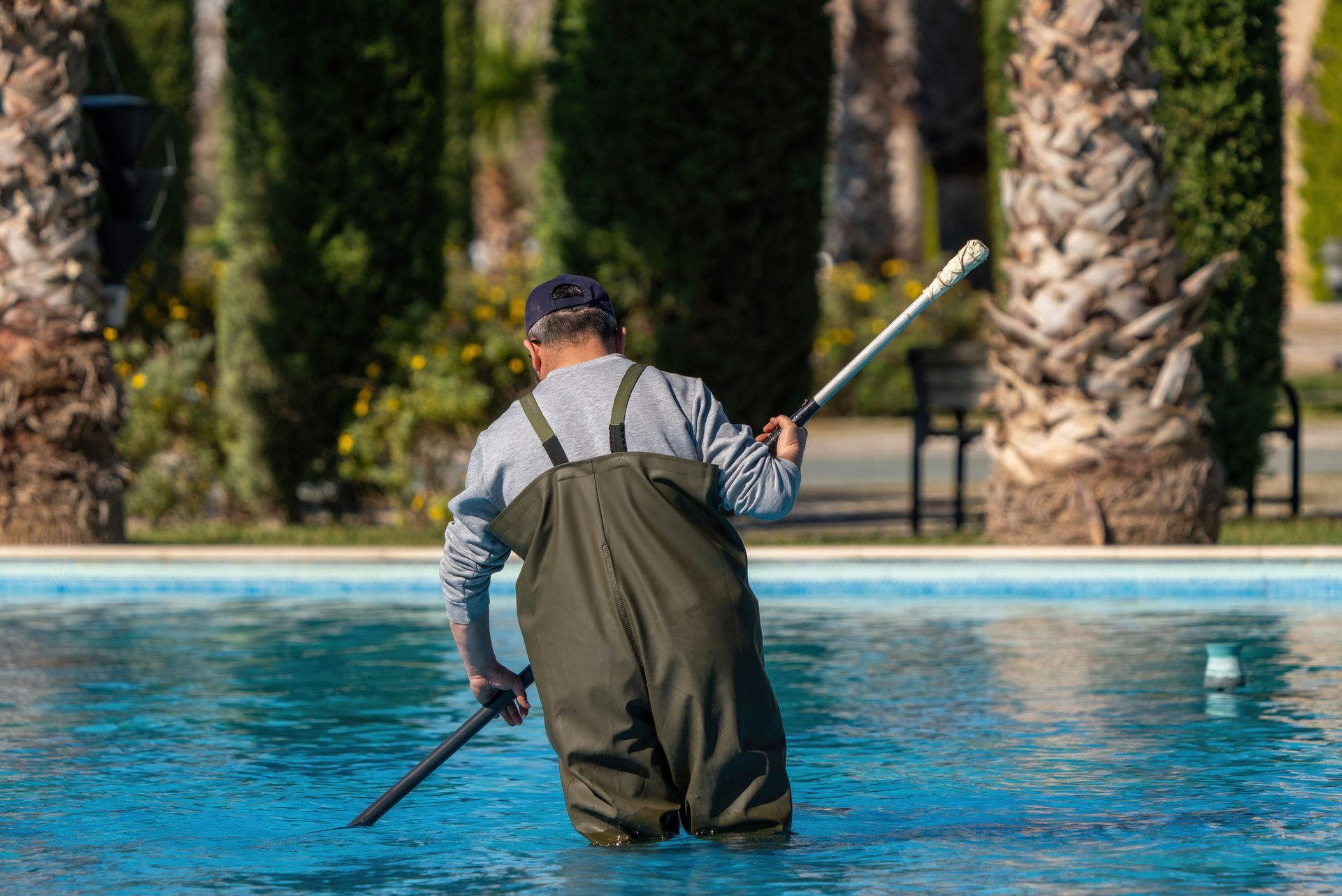 Pool technician in waders cleaning a swimming pool with a telescopic pole.