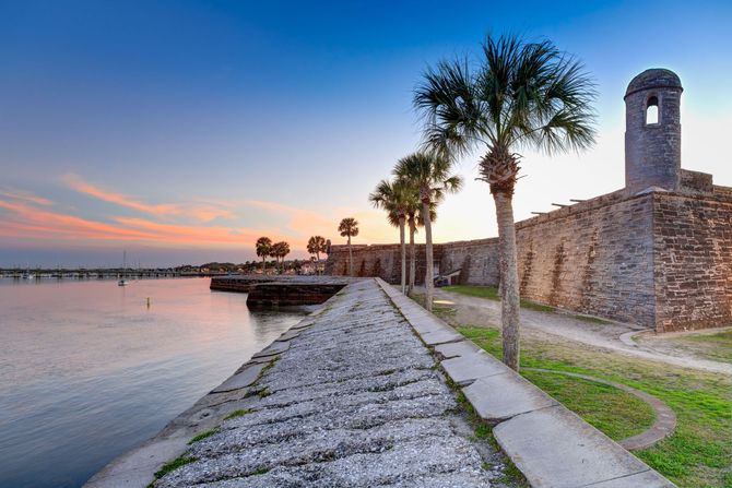 A stone walkway leading to a castle with palm trees and a sunset in the background.