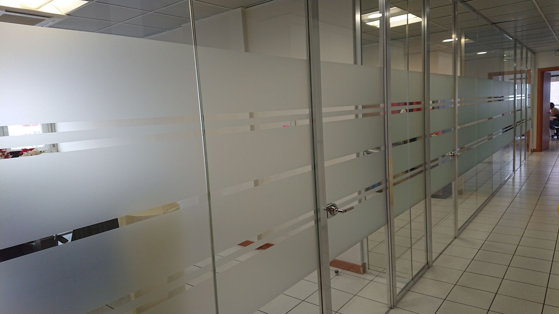Corridoio moderno per uffici con pareti divisorie in vetro satinato e pavimento piastrellato.