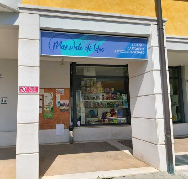 Un piccolo bar con vetrina a righe, ombrelloni da esterno, sedie e insegna a muro, situato in una strada tranquilla.