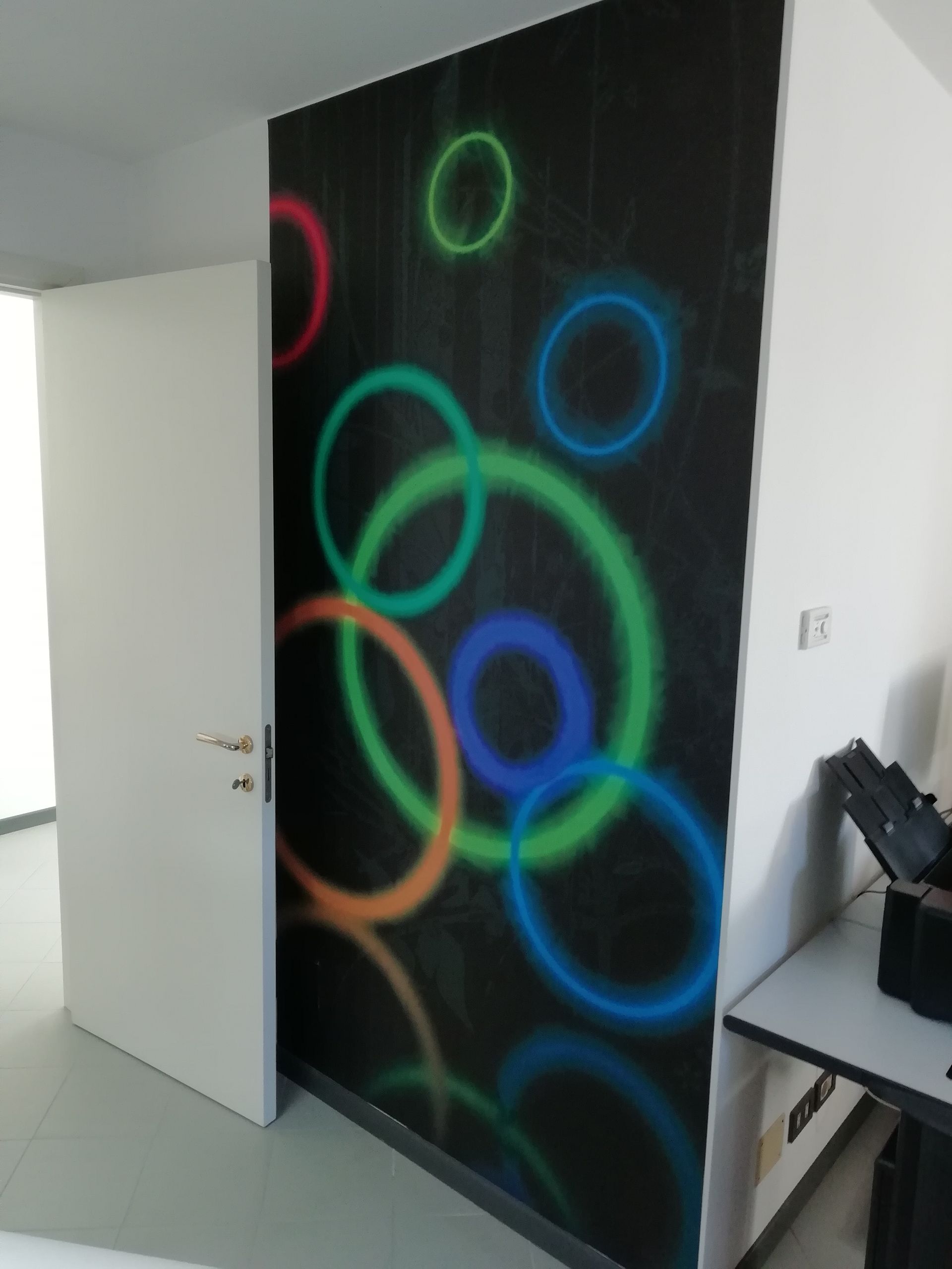 Un murale con anelli al neon colorati su una parete nera in una stanza bianca, con una porta aperta e una scrivania nelle vicinanze.