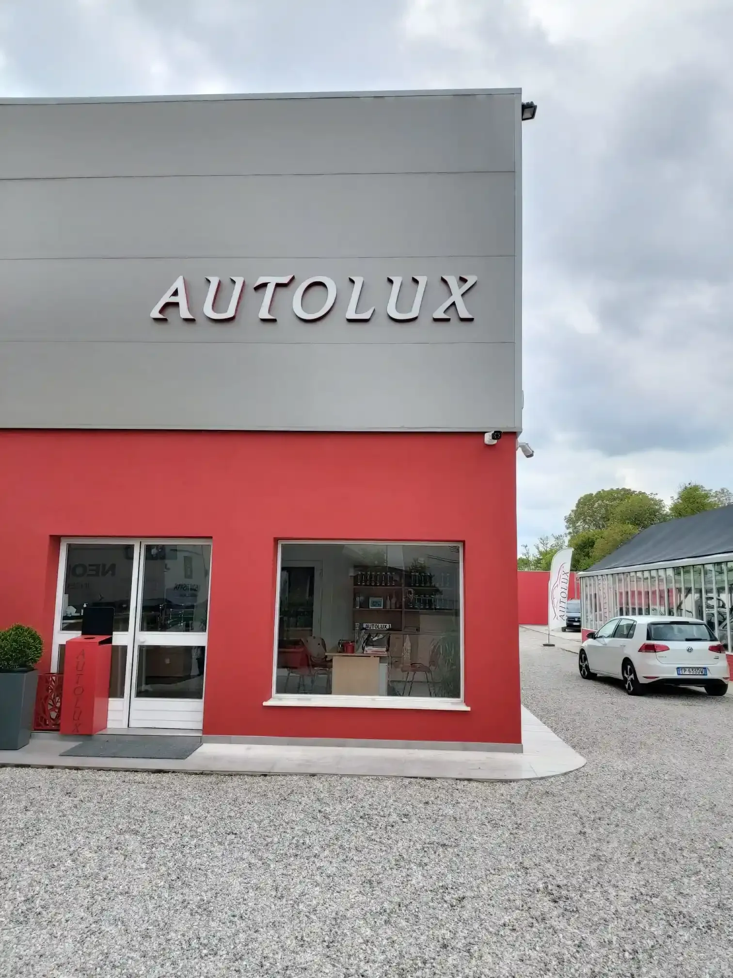 Vetrina del negozio AUTOLUX con parte inferiore della facciata rossa, parte superiore grigia e auto parcheggiate all'esterno.
