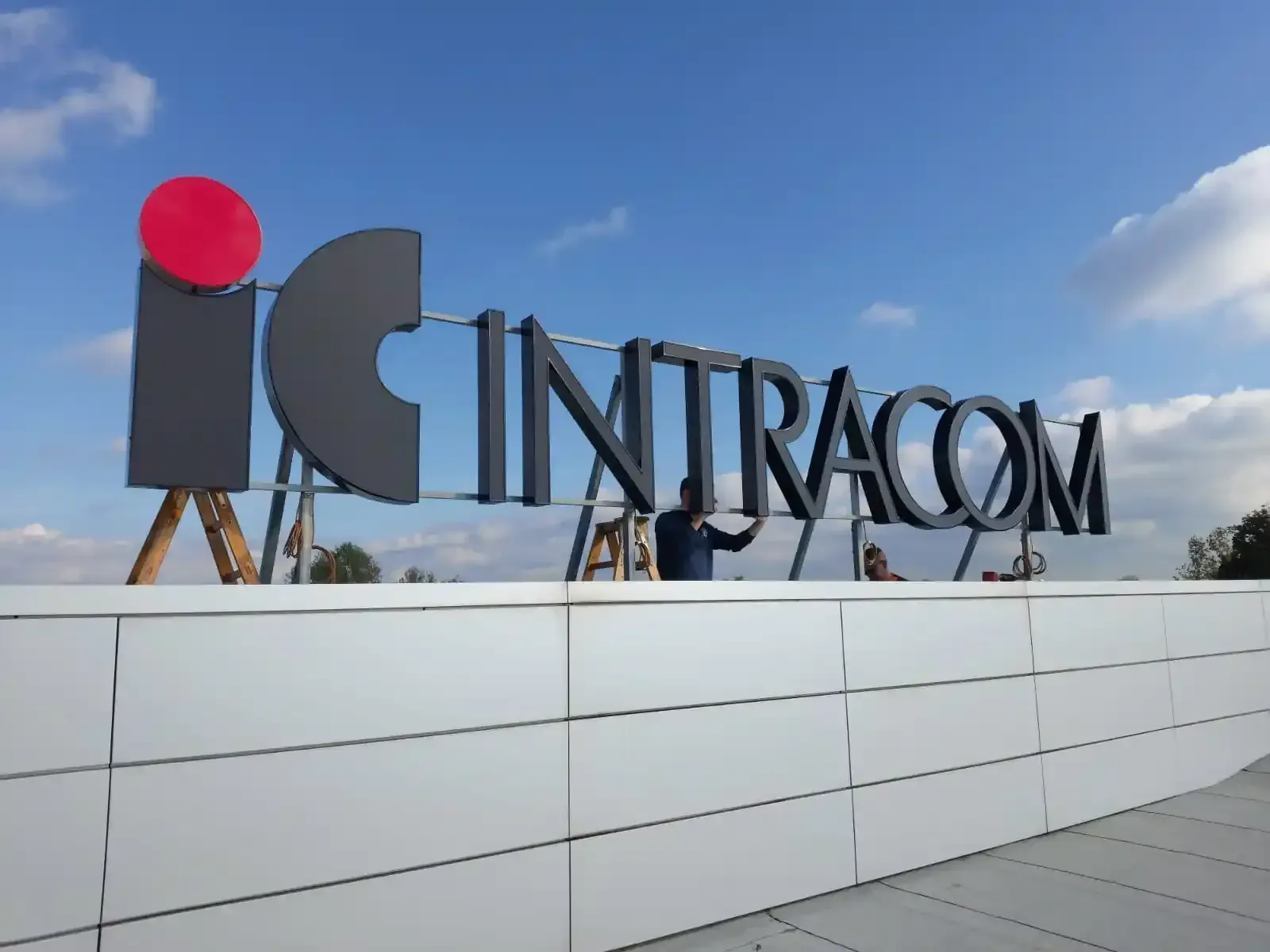 Insegna INTRACOM sul tetto, stagliata contro un cielo azzurro, con un muro bianco in primo piano.