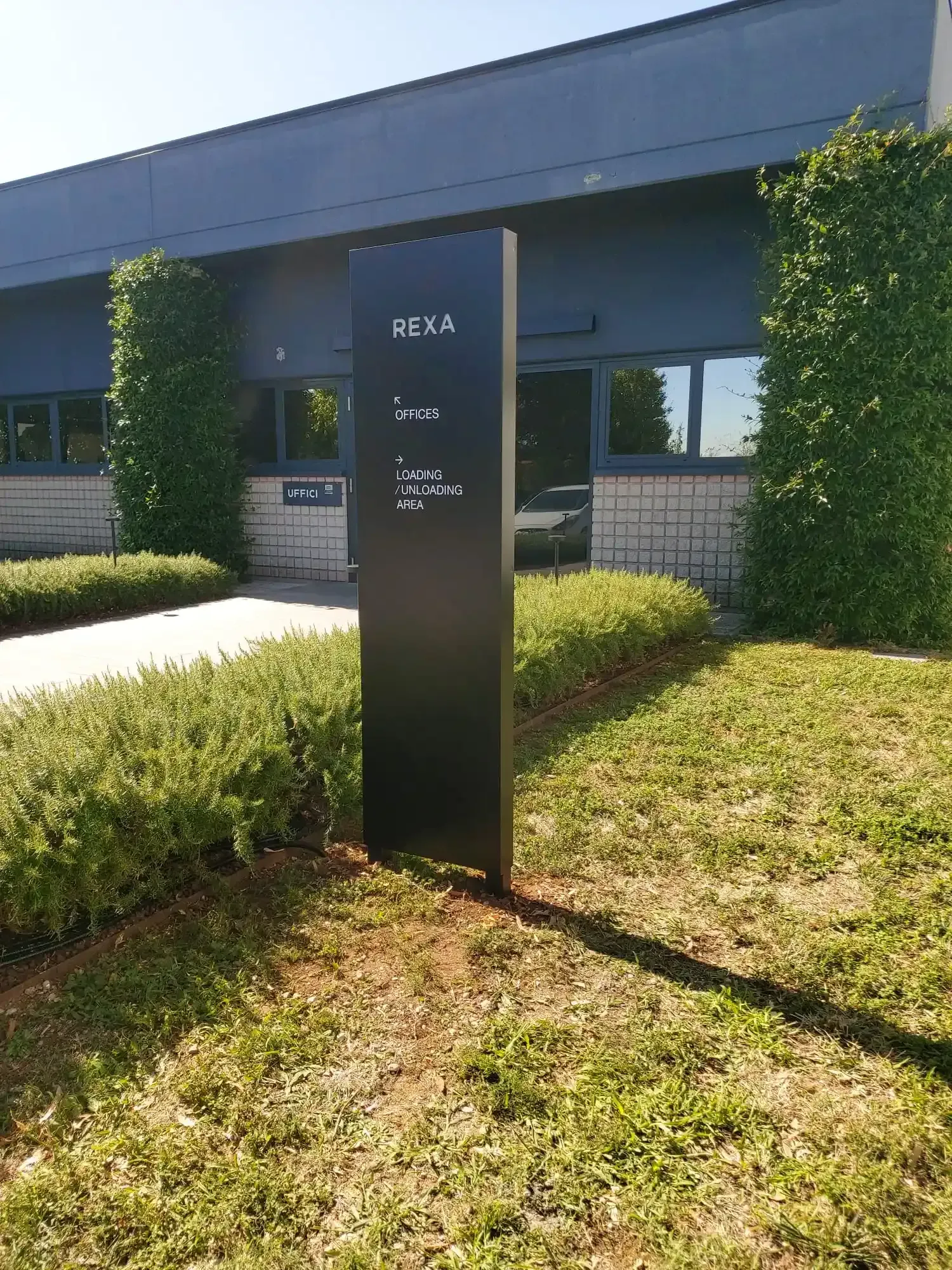Cartello nero all'esterno di un edificio moderno, incorniciato da arbusti verdi ed erba in piena luce solare.