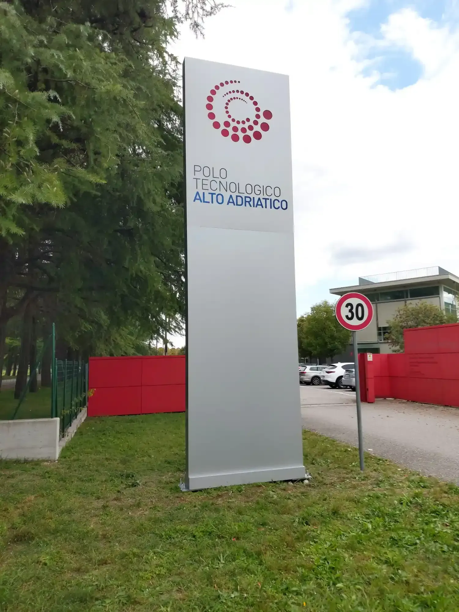 Alto cartello bianco per la Fundación Alto Adriático accanto al cartello stradale che indica la velocità di 30 km/h e al cancello rosso.