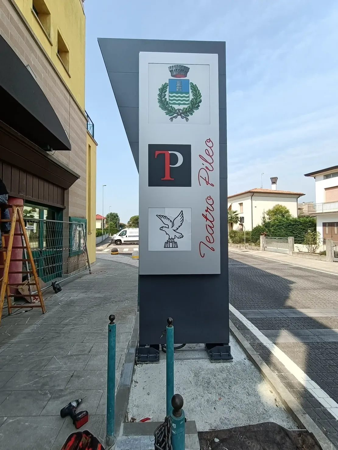 Segnale stradale con diversi loghi su un pannello grigio, a lato di una strada acciottolata e di alcuni edifici.