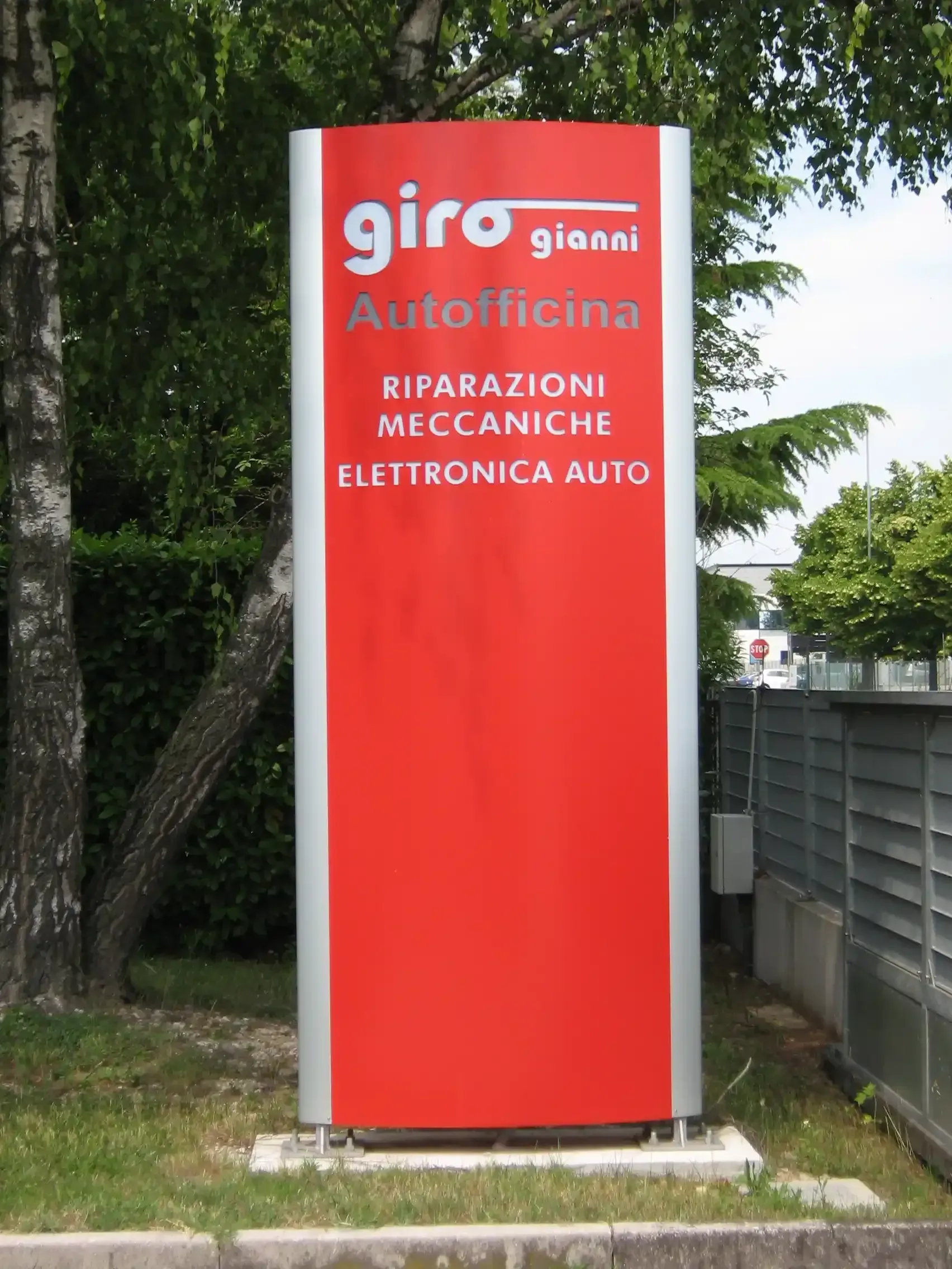 Insegna rossa esterna di un'officina meccanica, posizionata accanto ad alberi e una recinzione.