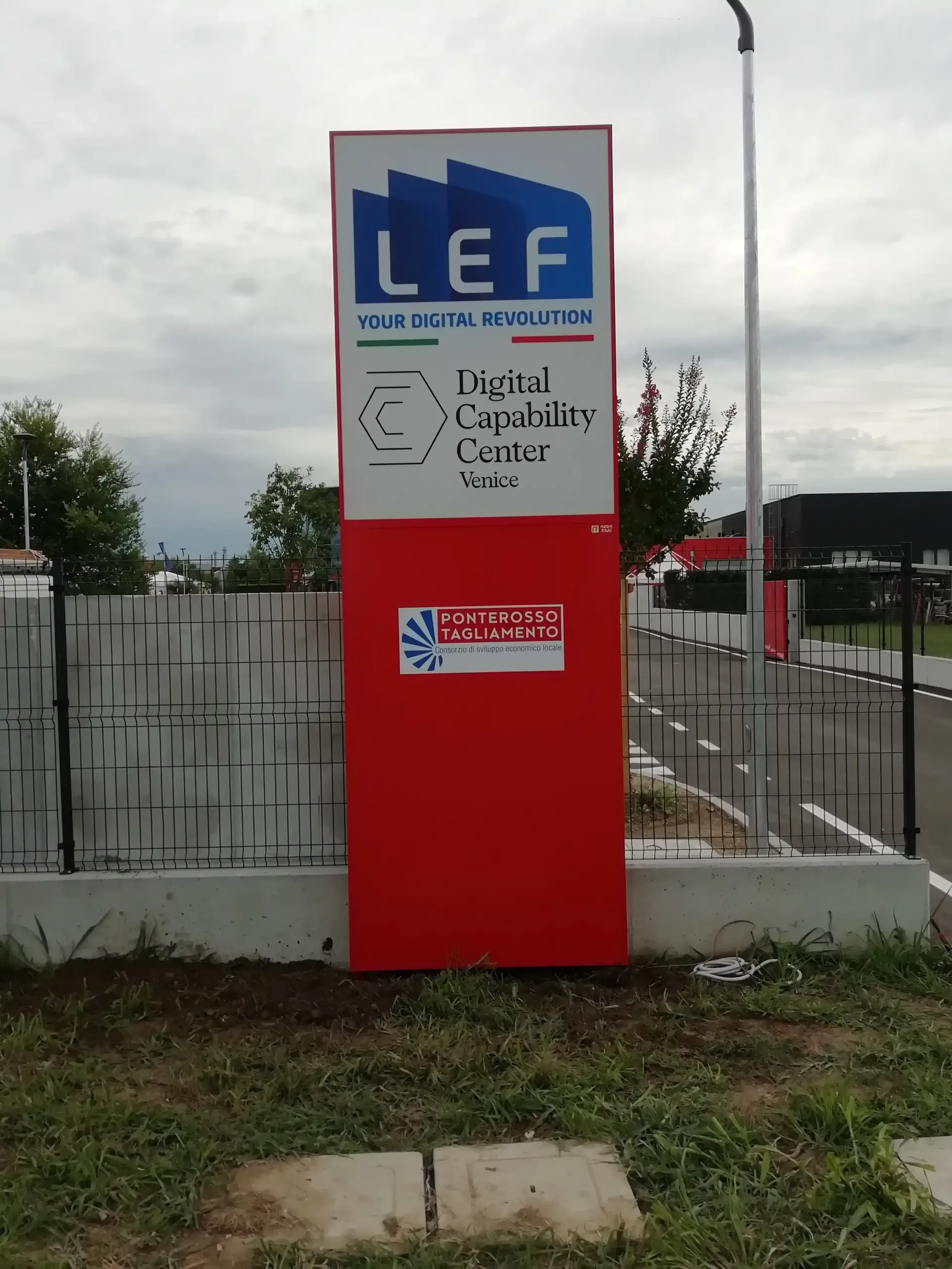 Cartello rosso LEF con pannello superiore bianco e avviso scritto a mano accanto a una recinzione a bordo strada.