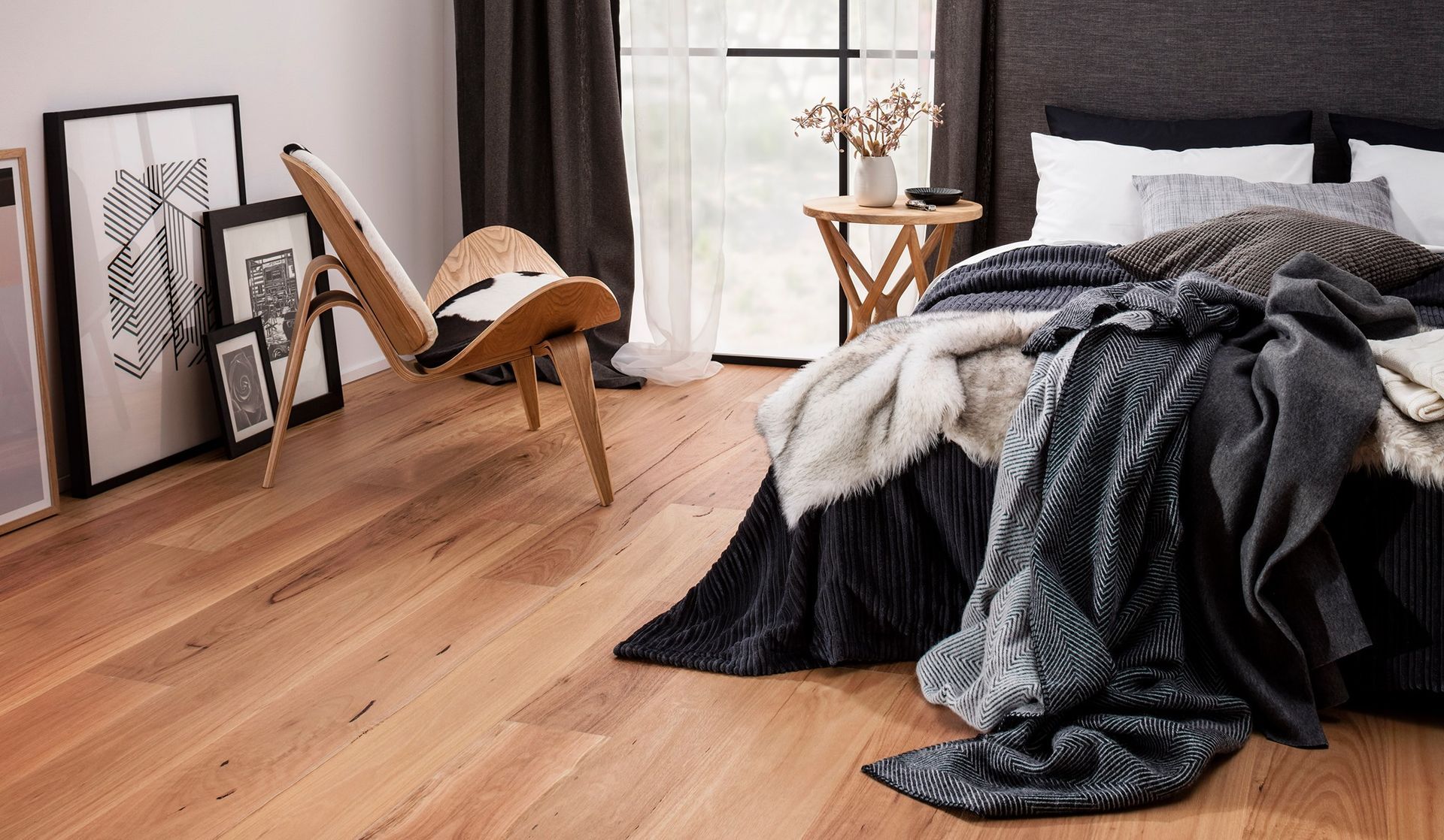 Bedroom with hardwood floor, bed with blankets, chair, framed art, and window with curtains.