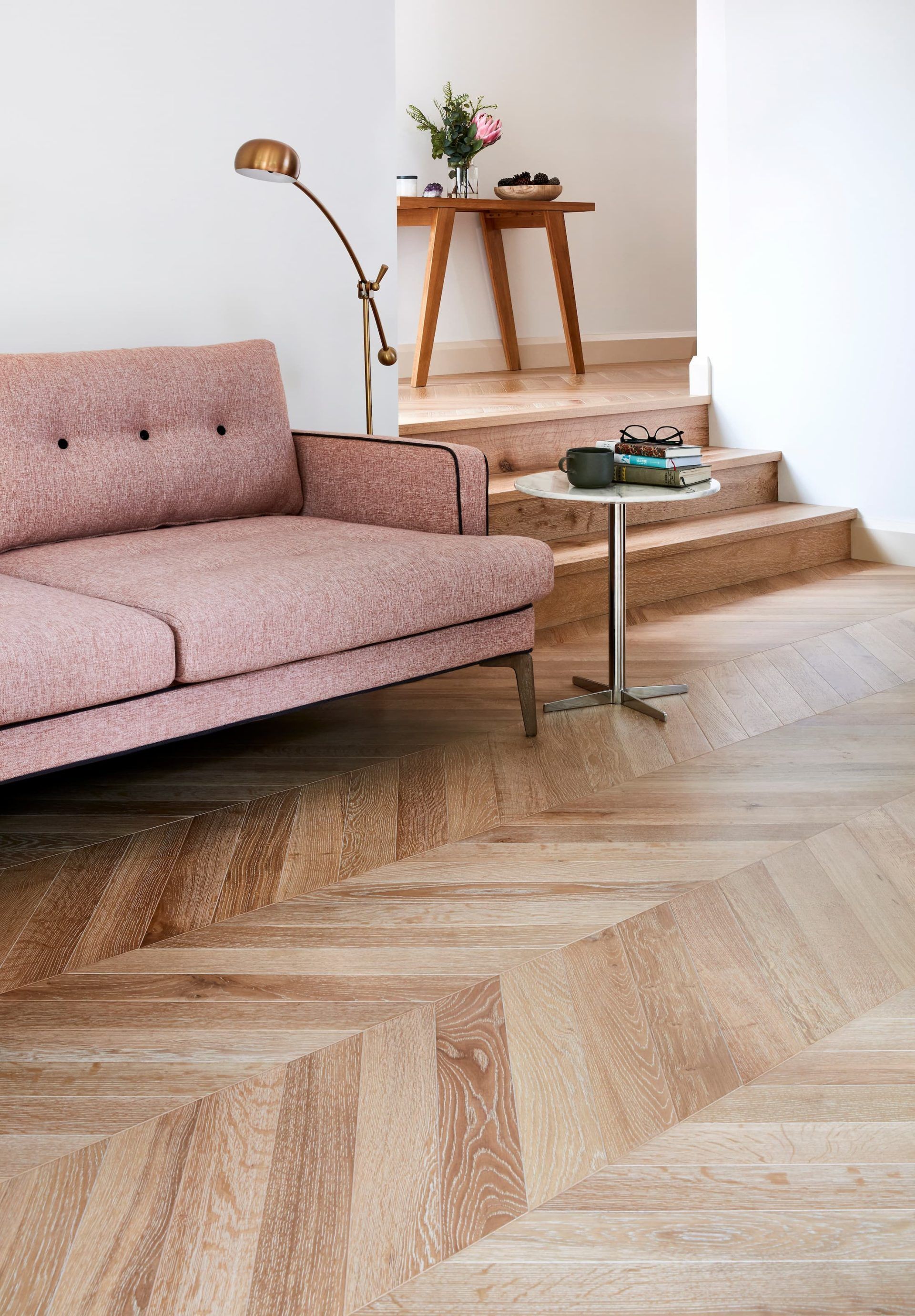 Pink sofa in a room with herringbone wood floor, lamp, side table, and small table with flowers.
