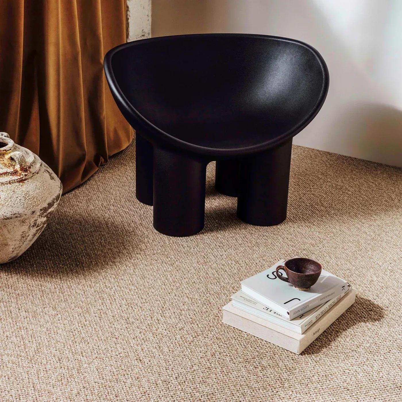 A dark, rounded chair with three legs sits on beige textured carpet, next to stacked books and a cup.