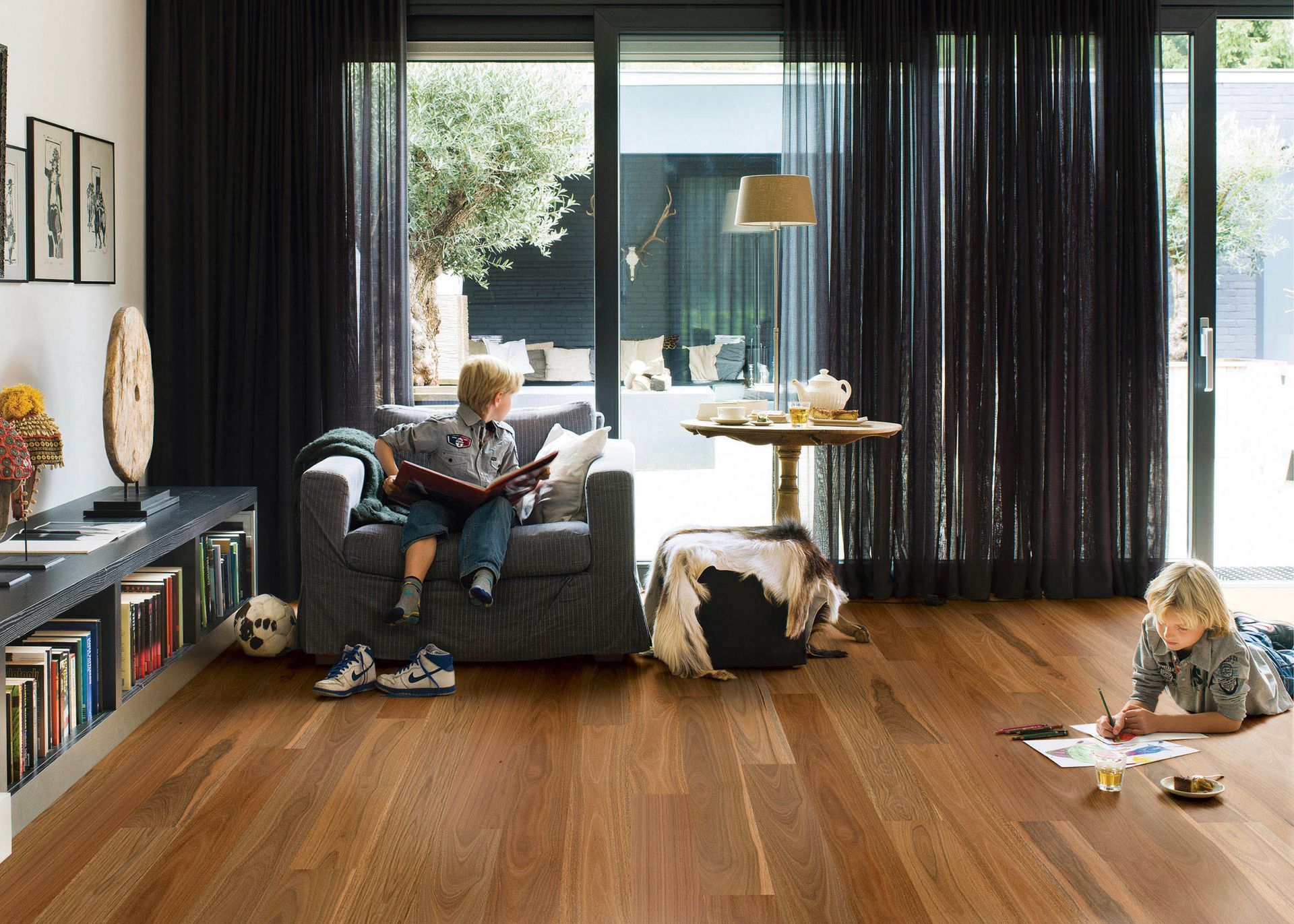 Two children in a sunlit living room with hardwood floors. One reads on a chair, the other draws on the floor. Black curtains frame the view.