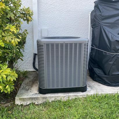 Outdoor air conditioner unit beside a house, partly covered by a black tarp, next to bushes and grass