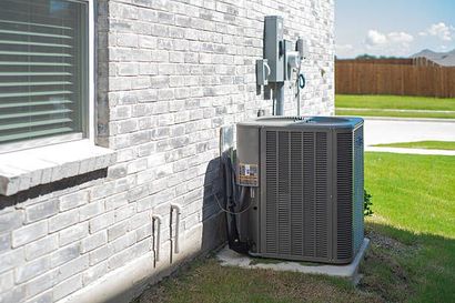 Outdoor air conditioner unit beside a white brick house and window on a sunny lawn