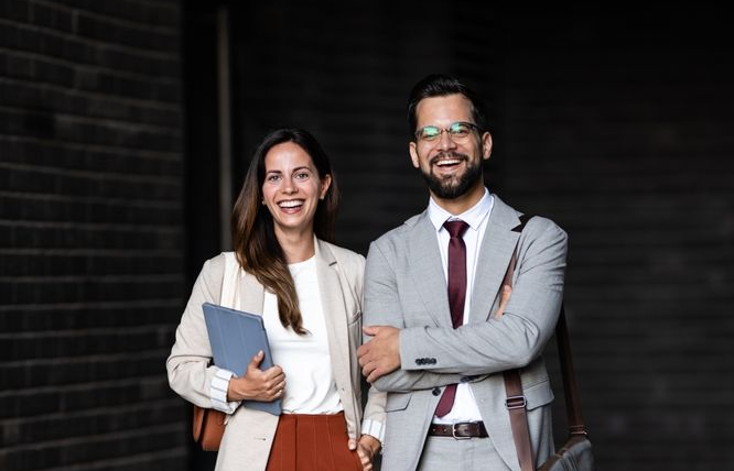 Woman and man in business attire smiling, standing in front of a dark brick wall.