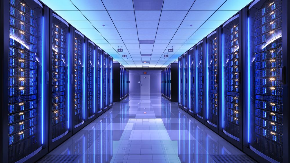 A corridor in a server room with rows of tall server racks illuminated by blue vertical lights, reflecting on the floor.
