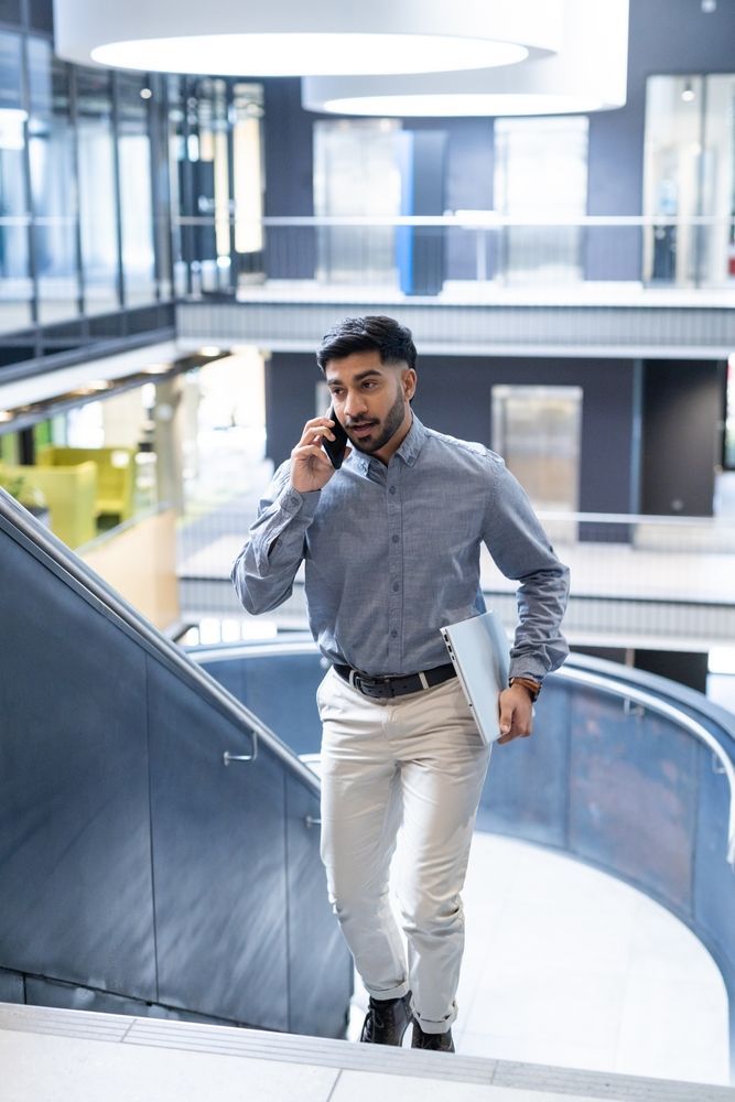 Business professional walking upstairs while on phone and carrying laptop in a modern office building.
