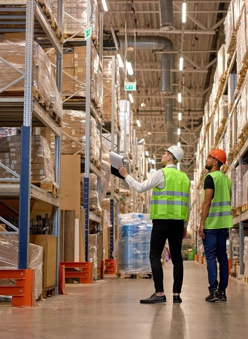 Two warehouse workers in safety vests inspecting shelves in a large storage aisle