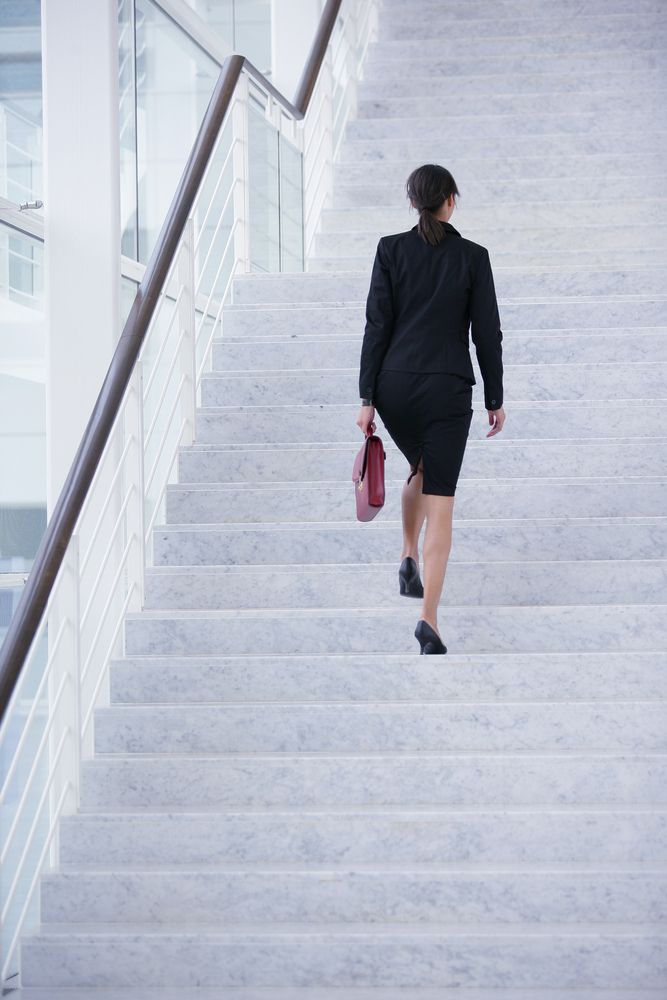 Woman in a business suit ascends a modern, light-colored staircase, holding a red briefcase.