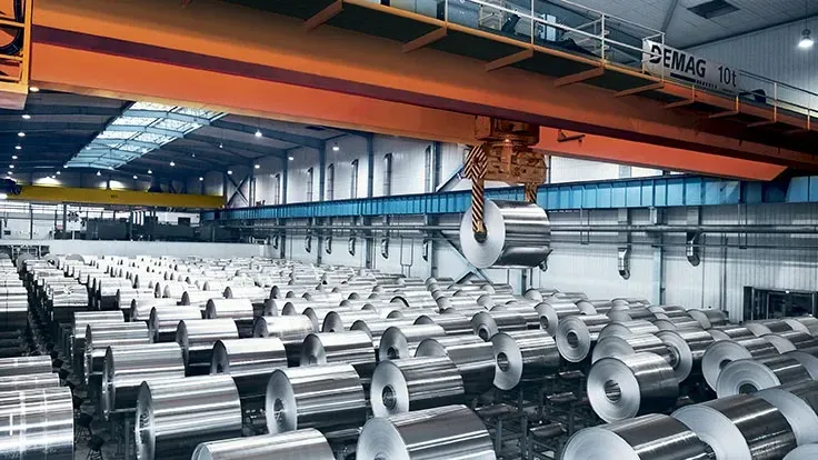 Industrial warehouse with rows of metal coils and overhead crane lifting steel, showing large-scale production.
