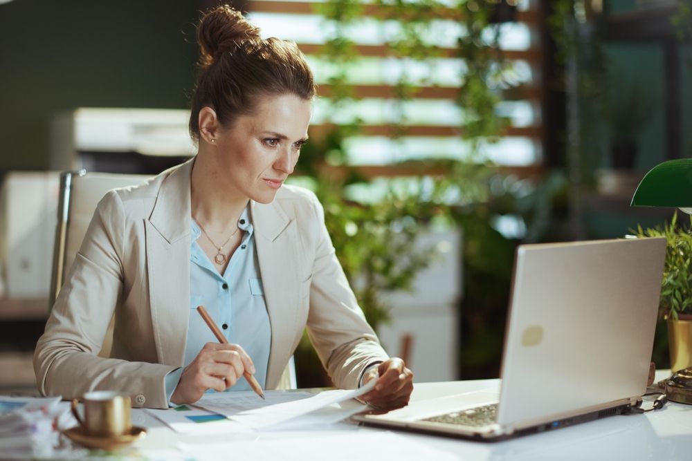 Woman in blazer working at desk, looking at papers and laptop.