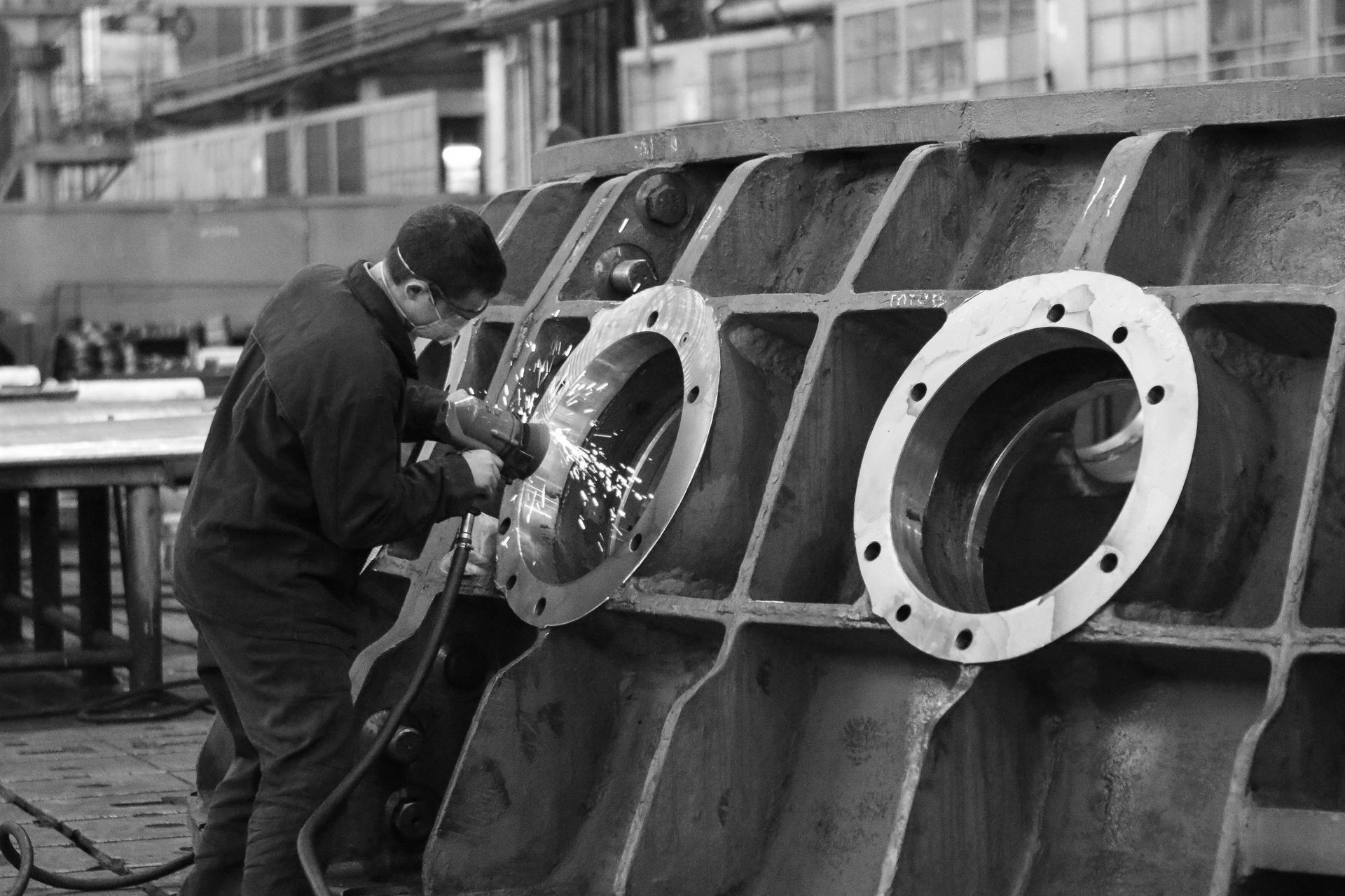 Factory worker grinding large metal component with sparks flying inside industrial manufacturing shop