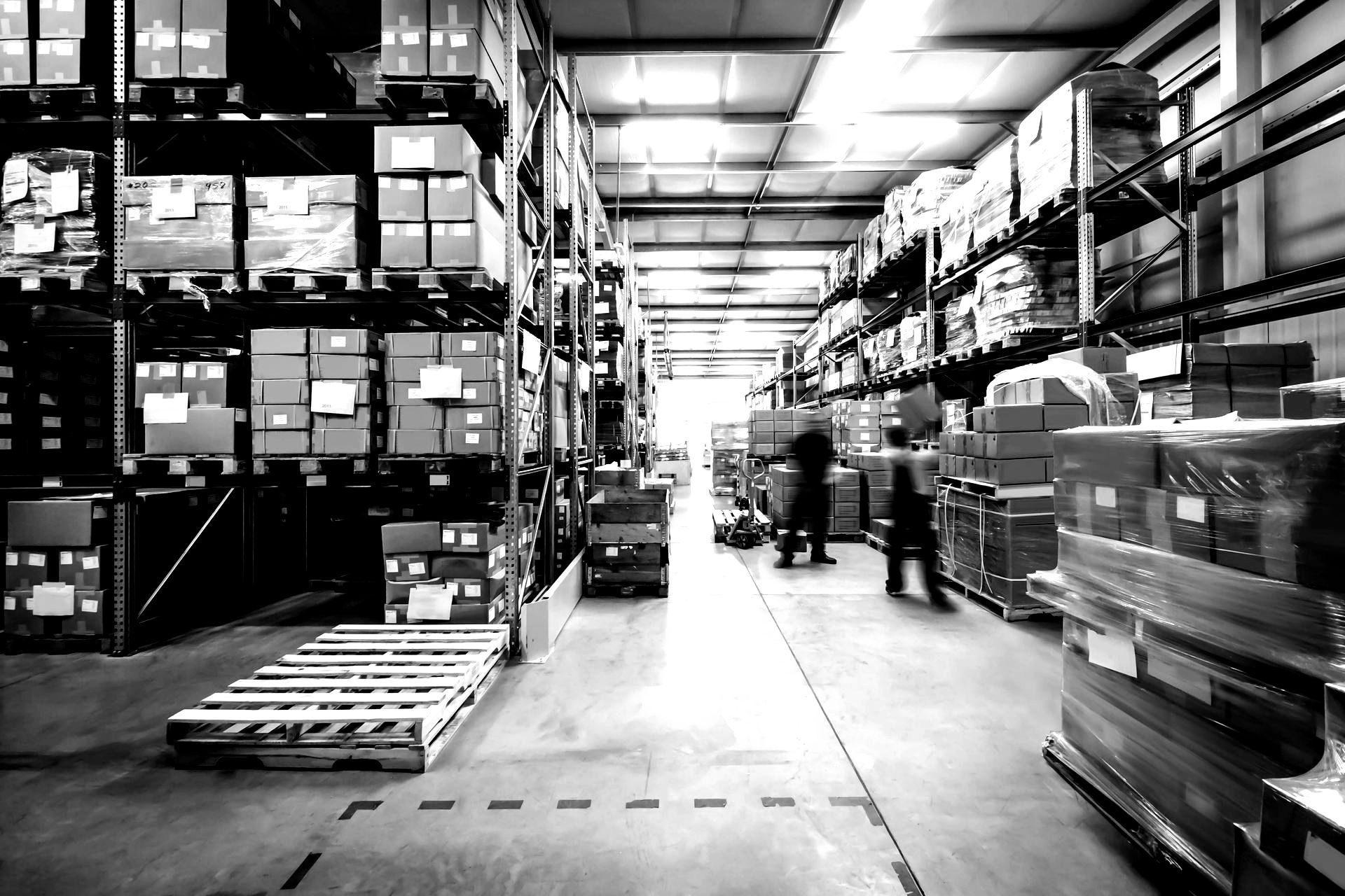Warehouse interior with tall shelving packed with boxes; workers near the center aisle.
