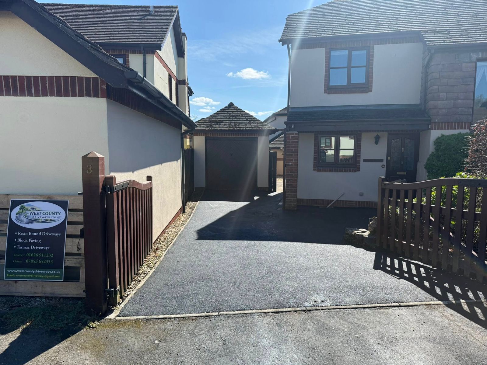 A sunny driveway leads between two houses toward a detached garage, with a sign displayed on a wooden fence to the left.