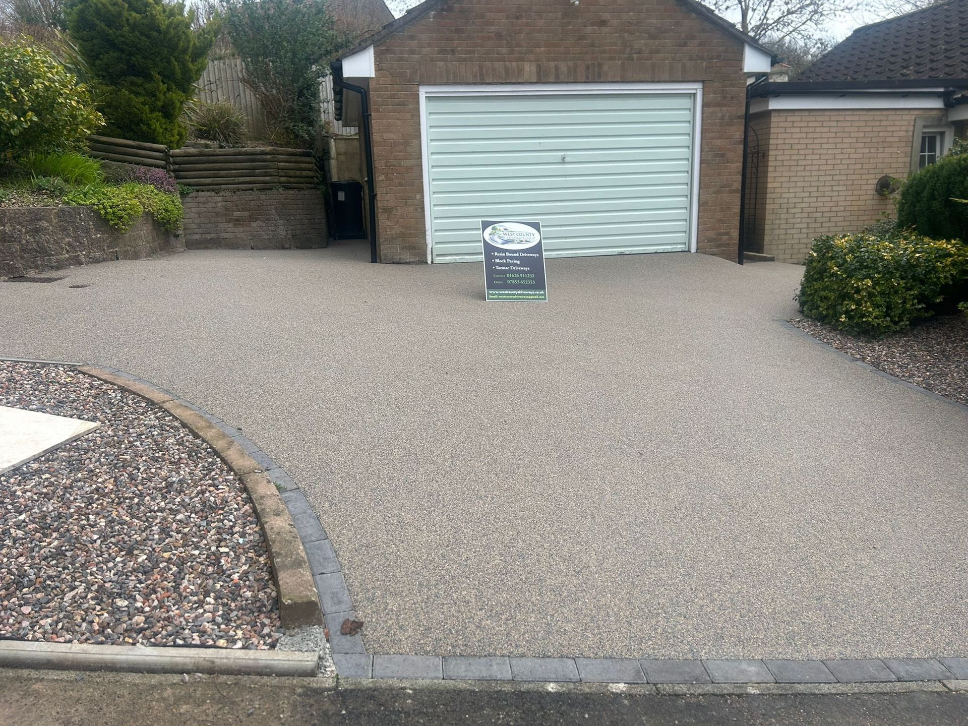 Gravel driveway leading to a white garage door, with a small sign centered in front and landscaped edges.