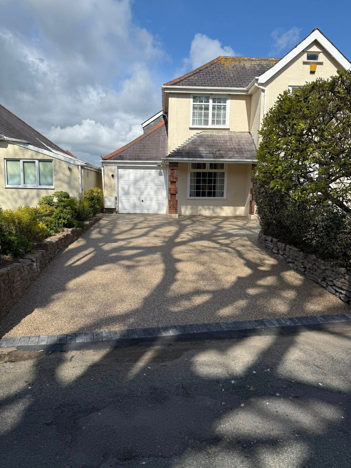 A light-colored two-story house with a white garage door and a gravel driveway partially shaded by tree shadows.