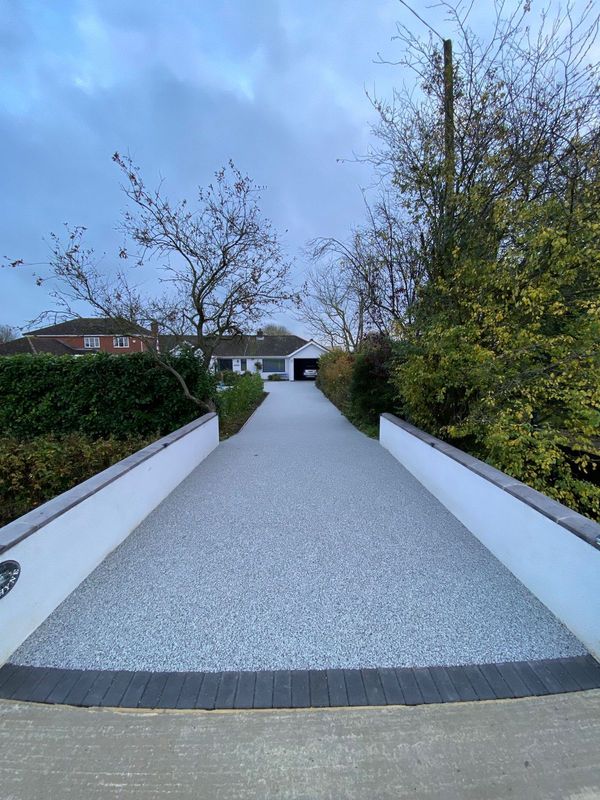 A long, grey resin driveway flanked by white walls and hedges, leading toward a house and garage under a cloudy sky.