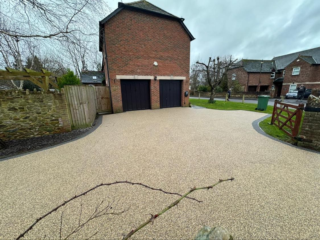 A paved driveway with light-colored aggregate, leading to a brick garage with two dark doors, set in a residential area.