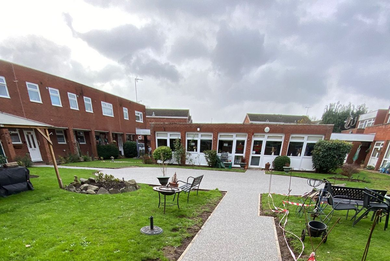 A courtyard with a stone path, grass, outdoor furniture, and red brick residential buildings under a cloudy sky.