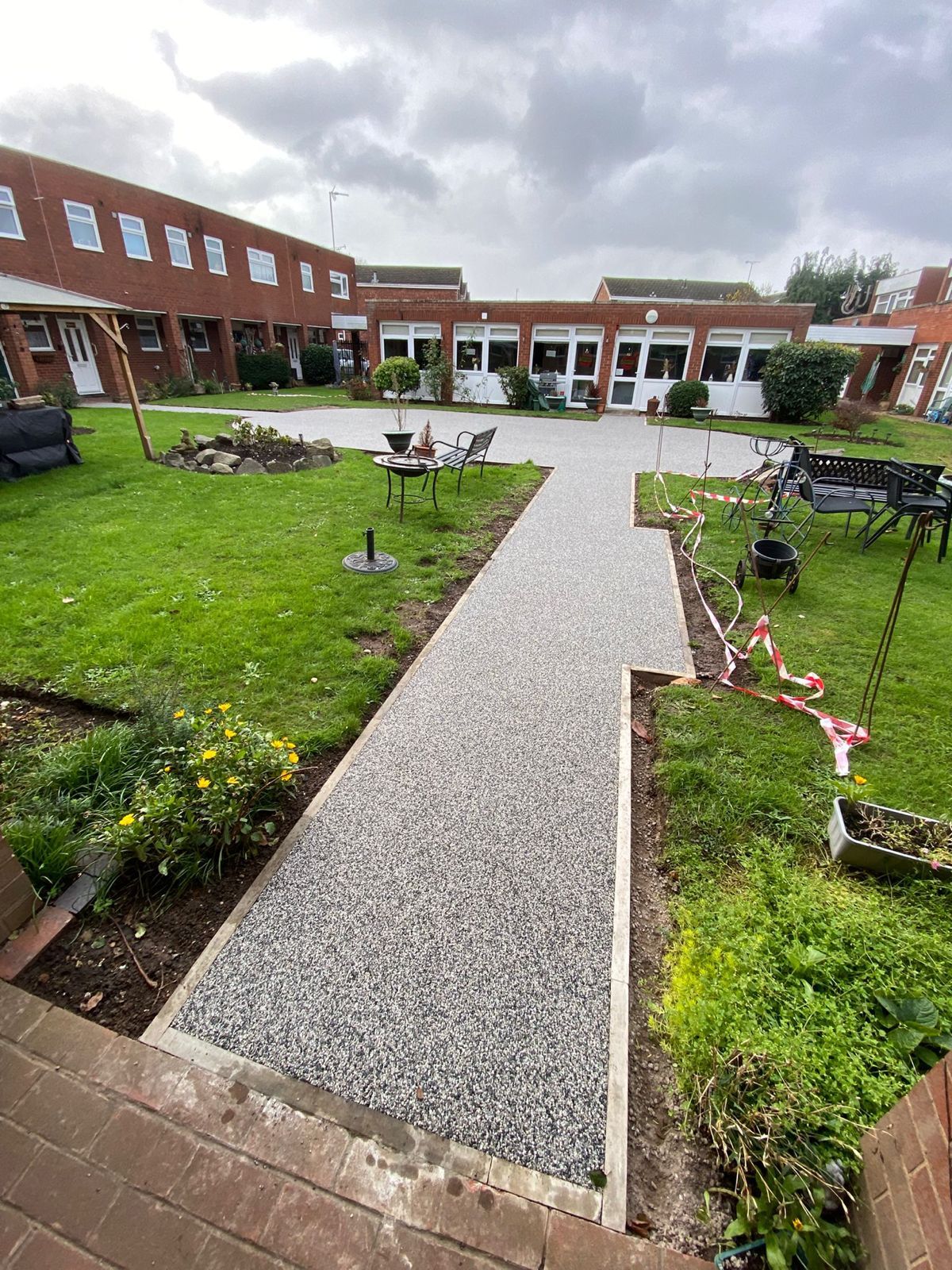A newly installed light gray gravel path runs through a residential lawn toward a red brick building complex under a sky.