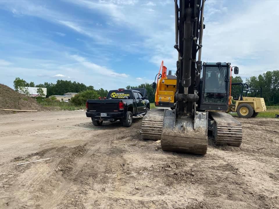 Black pickup truck and excavator on a dirt lot under a cloudy sky. A yellow machine is in the background.