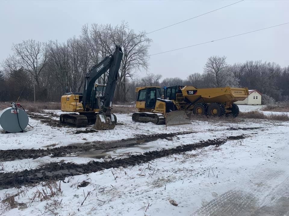 Excavator and dump truck in snowy field. Construction equipment near a fuel tank and trees.