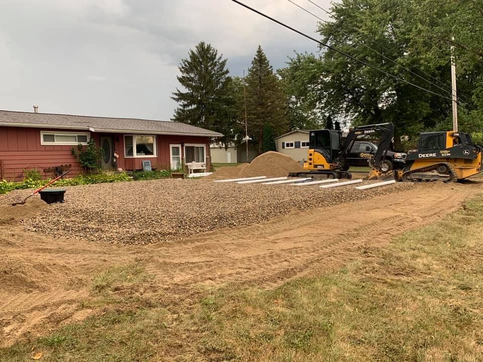 Construction site: two yellow tractors working near a red house, gravel and white pipes visible.