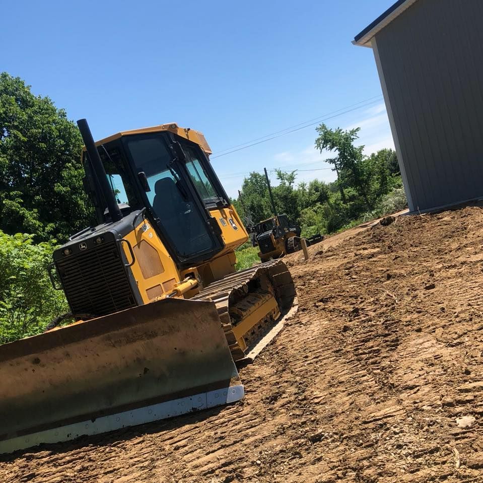 Yellow bulldozer on dirt terrain, clearing land near a building on a sunny day.