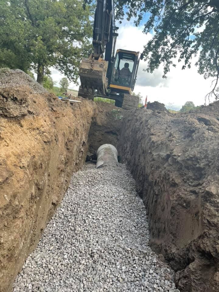 Excavator working in a trench, laying pipe. Gravel bed and soil walls. Green foliage in the background.