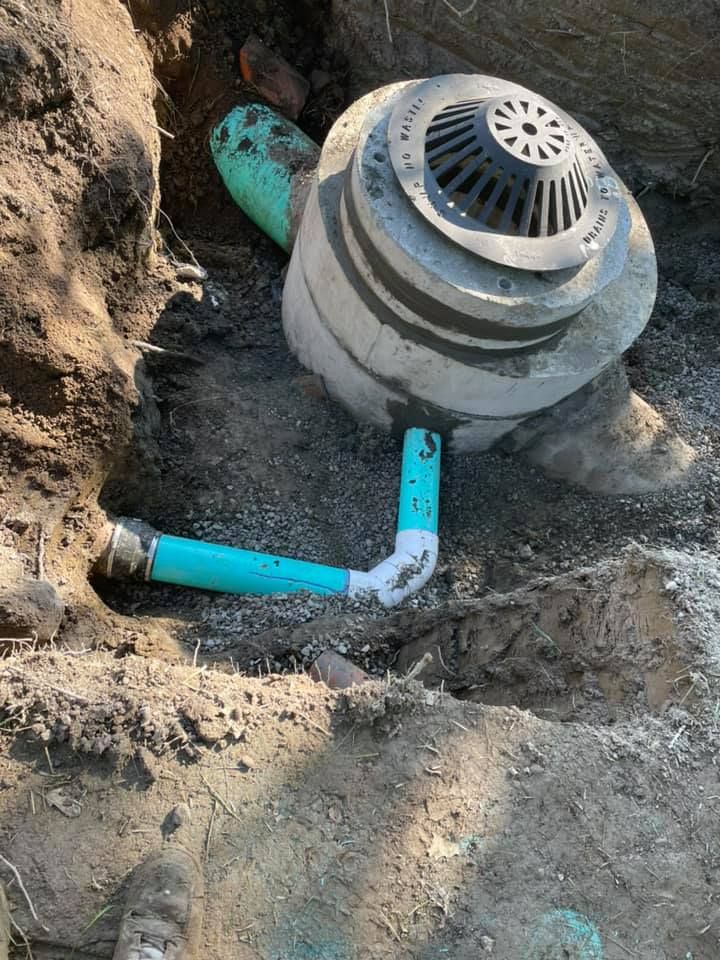 A concrete drainage catch basin with PVC pipes in a dirt trench, seen from above.