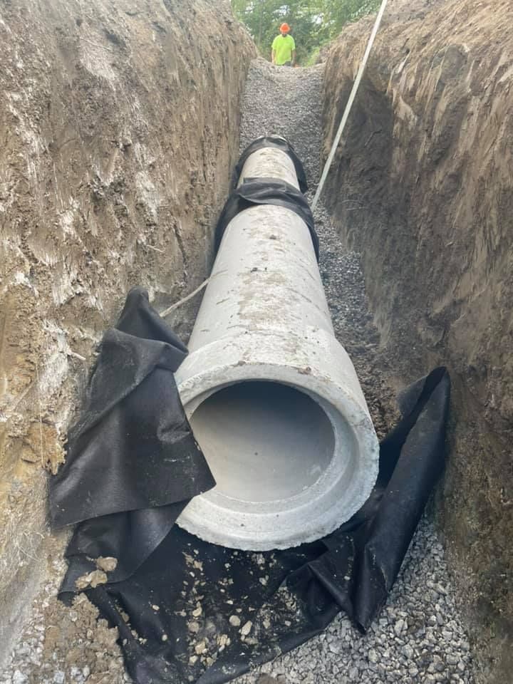 Concrete pipe in a trench, prepared for burial, with black fabric and gravel base. Person in yellow vest in background.
