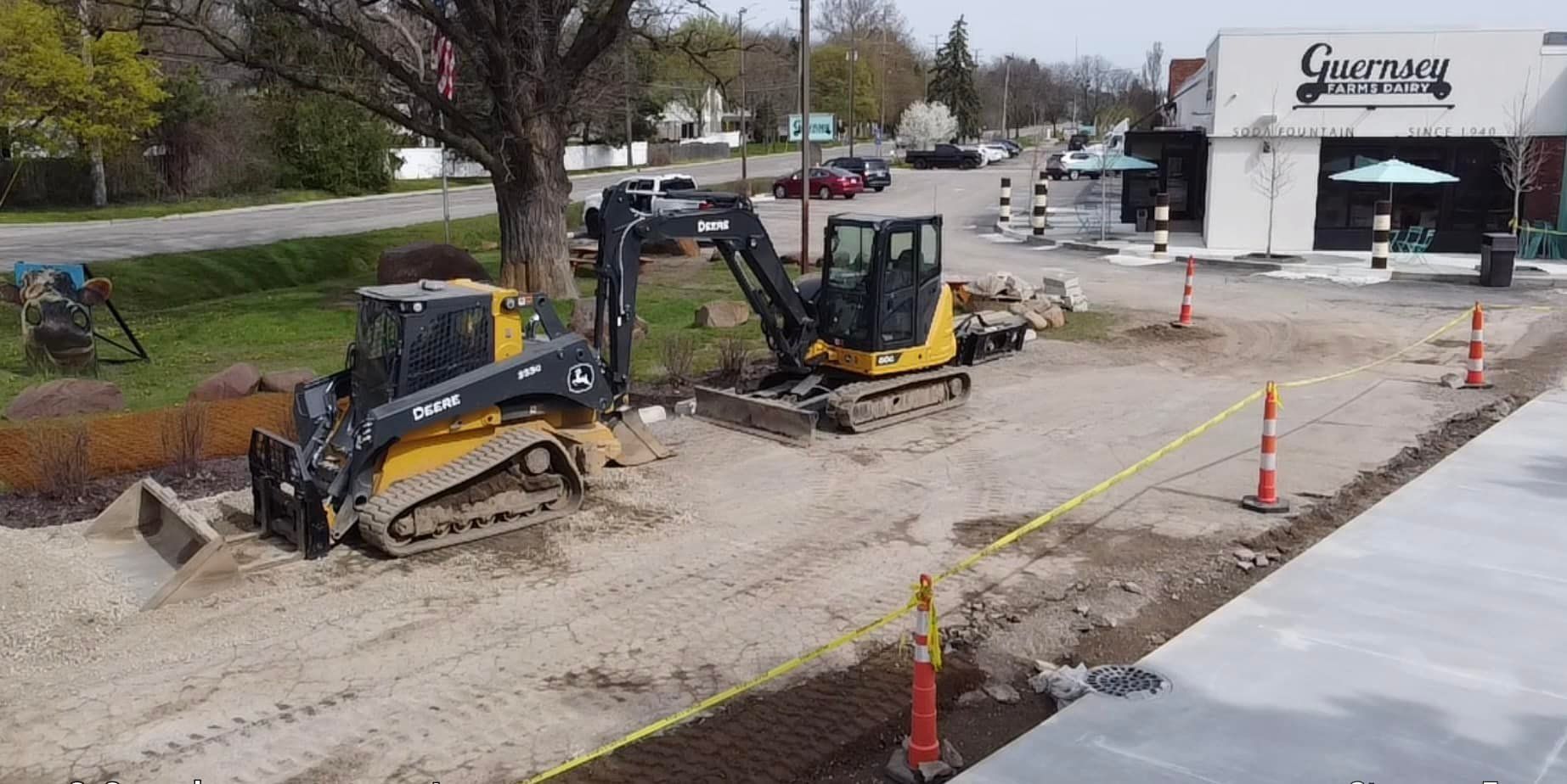 Construction site with two yellow excavators on a gravel road, near a tree and a building.