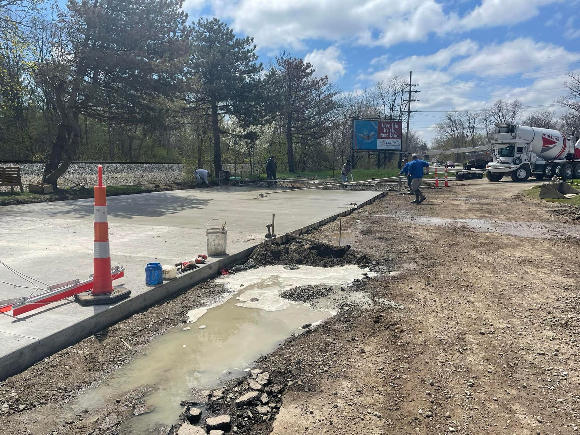 Construction site with a concrete pour. A worker and cement truck are visible. Orange and white traffic cones are present.